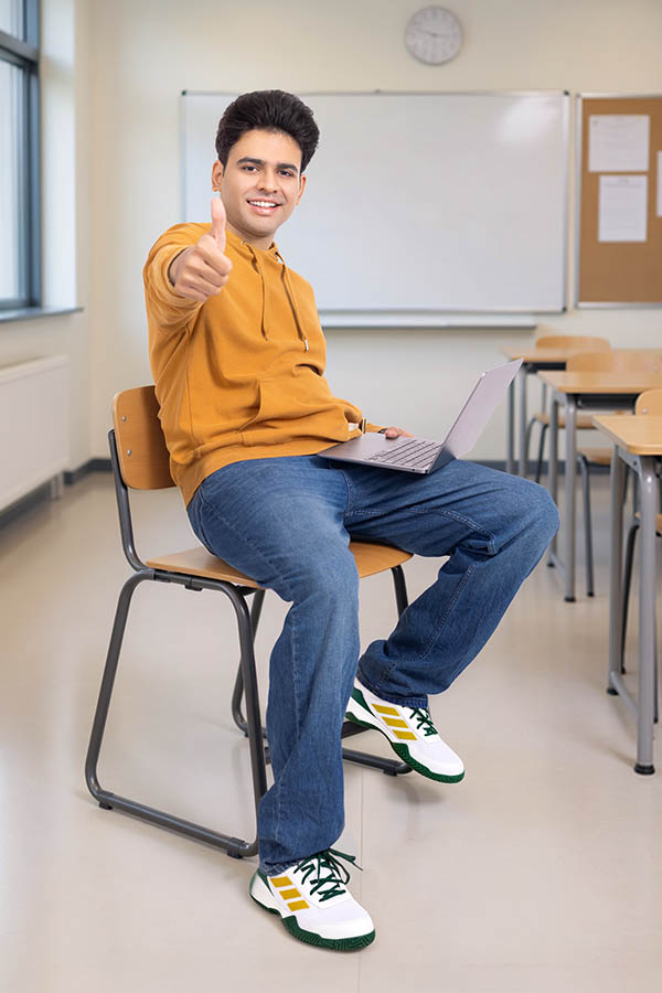 Young Indian male student showing thumbs up while using laptop in classroom