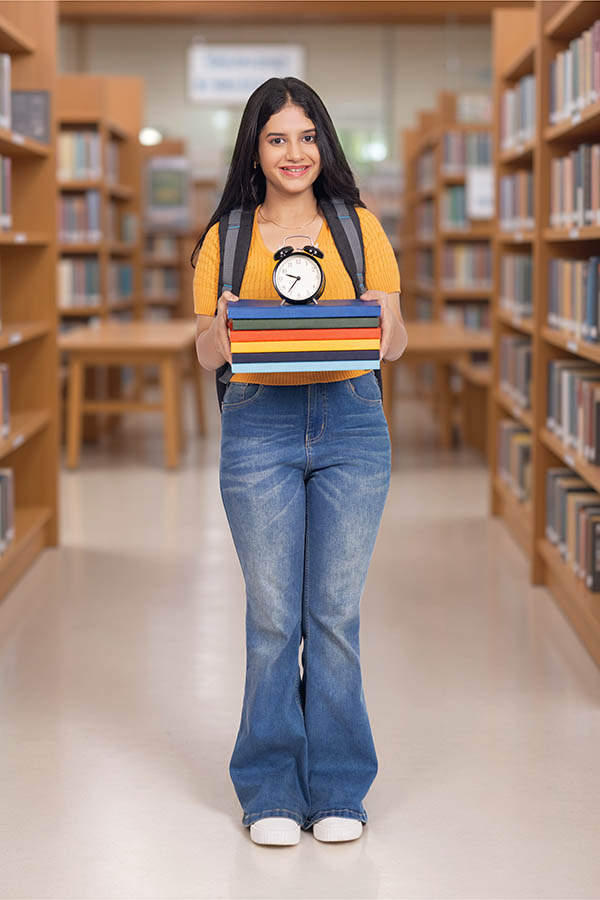 Indian college girl holding books and alarm clock in library