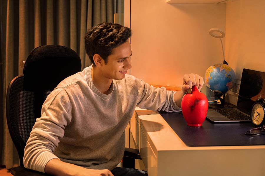 Young Indian student saving money in piggy bank at study desk