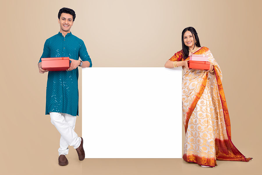 Indian couple holding gift boxes beside blank display board during festive celebration