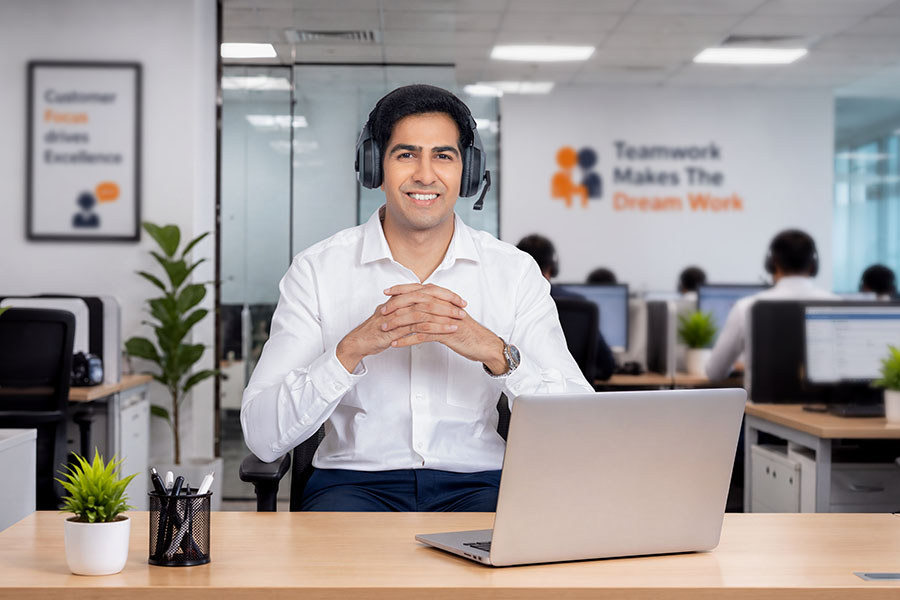 Smiling male customer service professional wearing headset with hands clasped at office desk