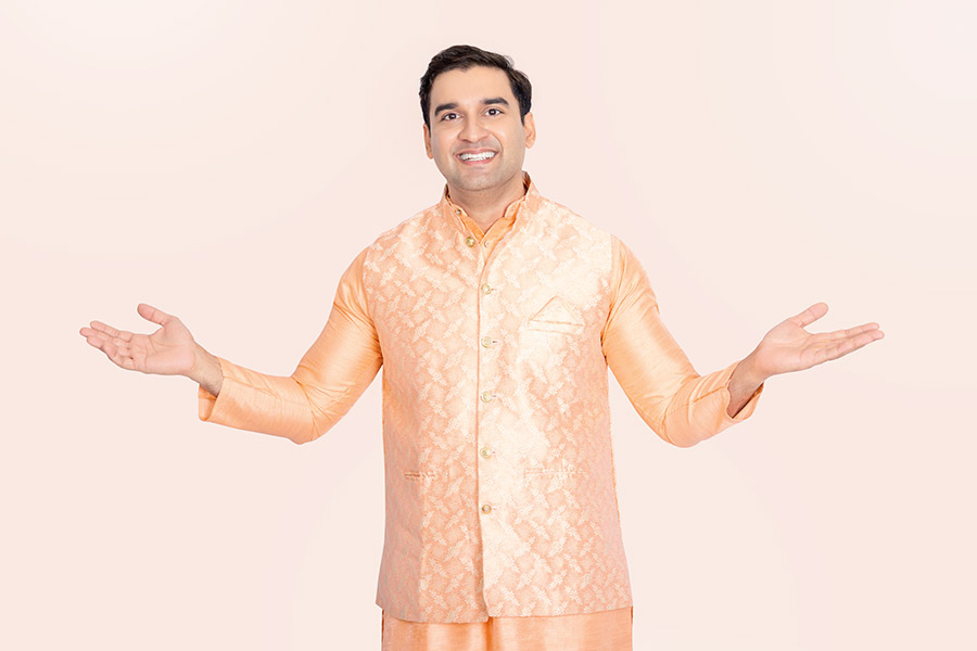 Indian man welcoming viewers in traditional peach attire against a pink background